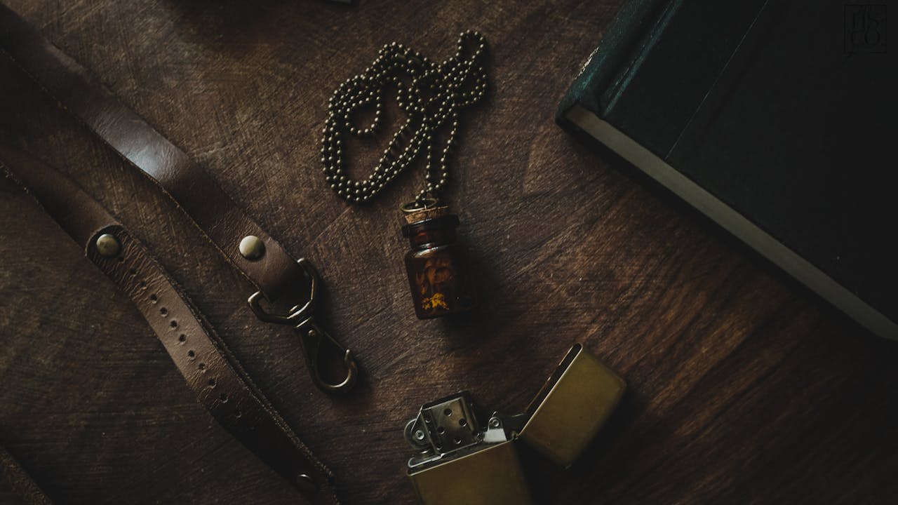 A rustic still life featuring a vintage vial necklace, lighter, and leather strap on a wooden table.